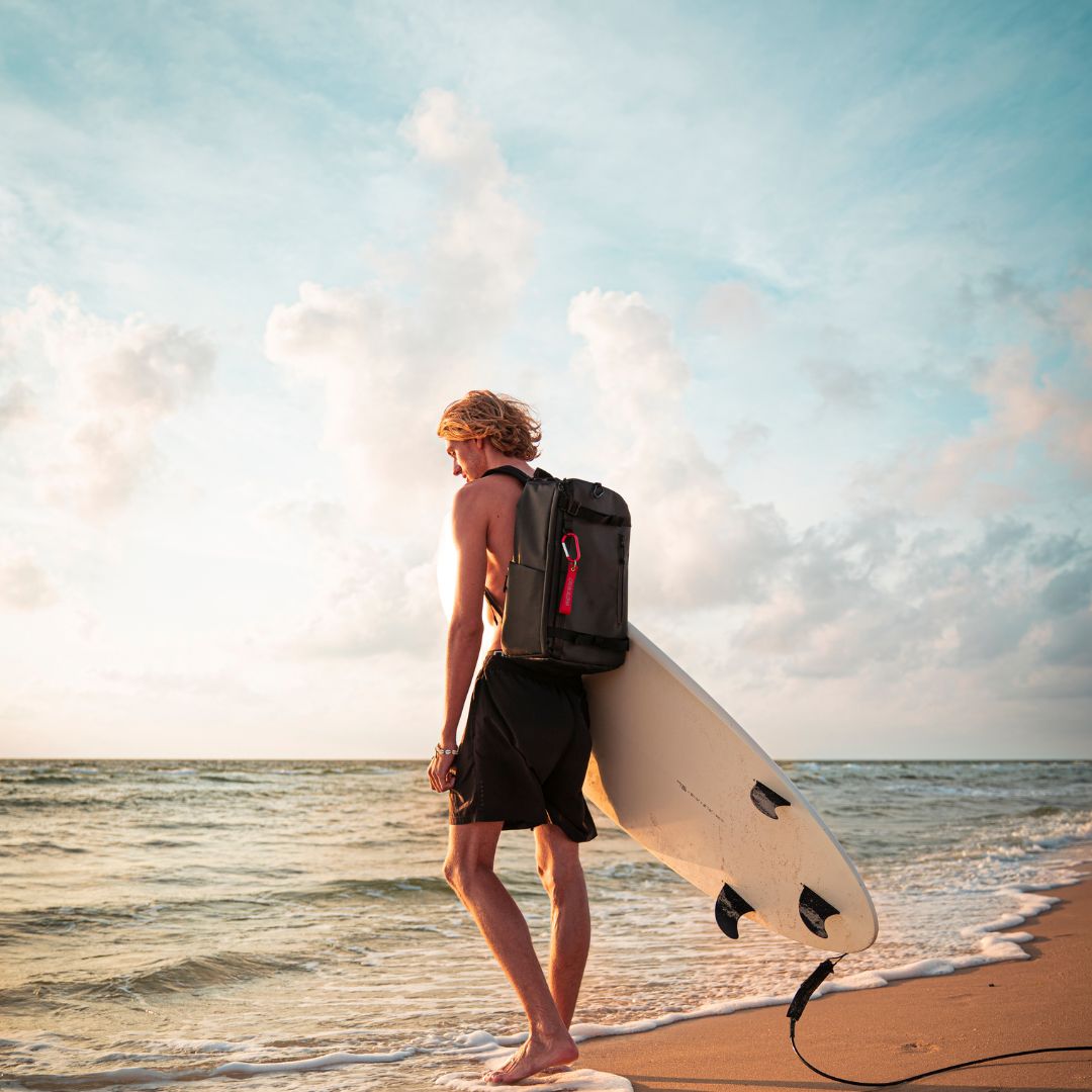 sons of aloha Outdoor Mann steht am Strand mit einem Surfbrett in der Hand und einem Rucksack auf