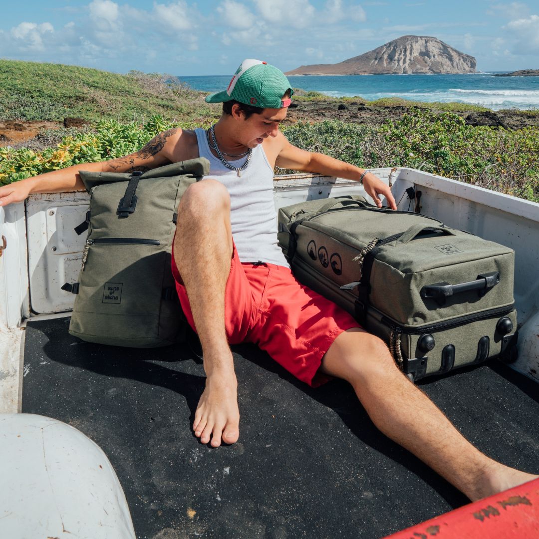 Outdoor Mann sitzt auf einer Ladefläche von einem Truck am Meer und neben sich der Rollkoffer mit Rollen in gün