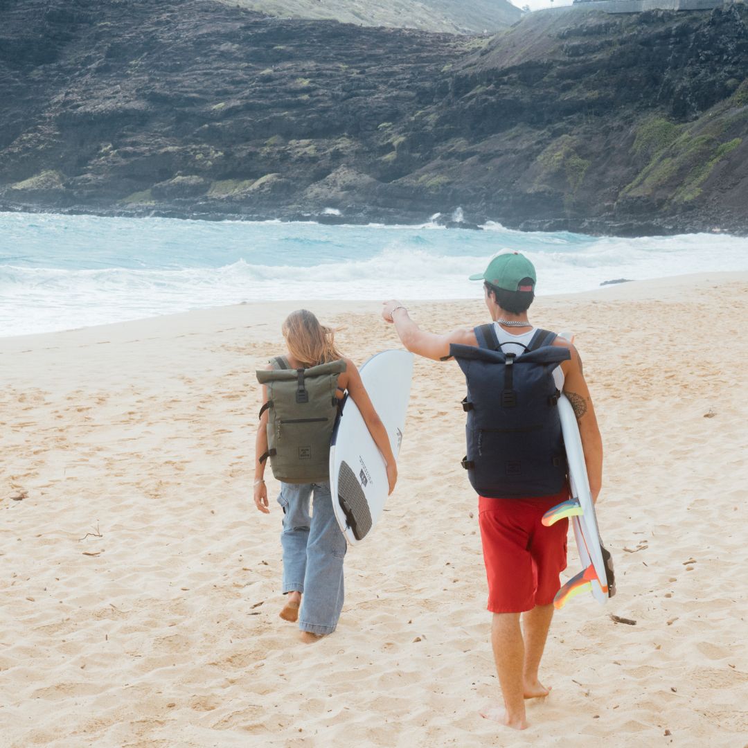 Rolltop Rucksack XL in grün und dunkel blau Outdoor Mann und Frau tragen ein Surfbrett und gehen am Strand.