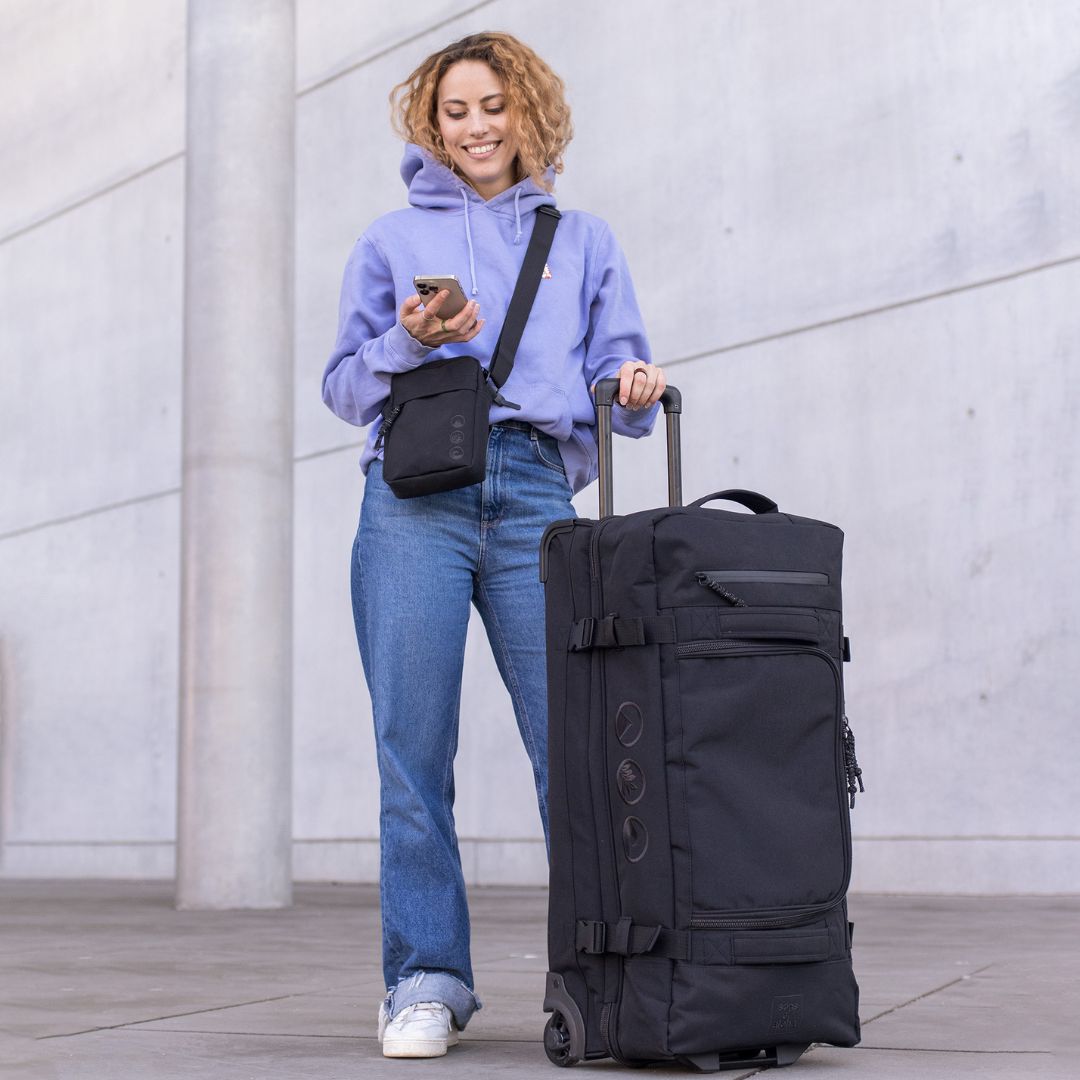Outdoor Frau traegt einen lila Hoodie und eine blaue Jeans neben ihr steht der schwarze Rollkoffer